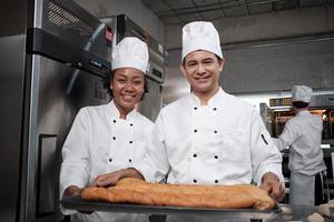 portrait de chefs professionnels en uniforme blanc regardant la caméra avec un sourire joyeux et fier avec un plateau de baguette dans la cuisine. ami et partenaire de la pâtisserie et de l'occupation quotidienne de la boulangerie fraîche. photo