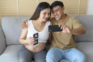 jeune femme enceinte avec mari embrassant et appel vidéo avec la famille et les amis par smartphone sur les médias sociaux, la famille et le concept de soins de grossesse photo