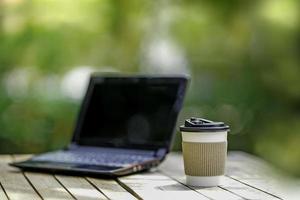 papier de tasse à café avec un ordinateur portable sur fond de nature verdoyante. travailler à distance ou à domicile. mise au point douce. effet de mise au point peu profonde. photo