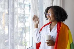 fille afro-américaine tenant un drapeau arc-en-ciel lgbtq dans sa chambre pour sortir du placard et célébrer le mois de la fierté pour promouvoir la diversité sexuelle et l'égalité dans le concept d'orientation homosexuelle photo