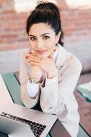 jeune femme d'affaires brune aux yeux charmants, mains douces avec manucure rouge portant une montre à portée de main et manteau blanc tenant les mains sous le menton assise près de son ordinateur portable se reposant après un travail acharné photo