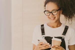 adorable fille teeange aux cheveux bouclés, vérifie le fil d'actualités, les messages sur les réseaux sociaux, utilise une connexion Internet gratuite, porte des lunettes optiques, un t-shirt et une salopette blancs décontractés, boit du café à emporter photo