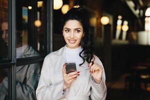 portrait horizontal d'une adorable femme brune en vêtements blancs utilisant son téléphone portable tout en se tenant à l'extérieur ayant de la bonne humeur touchant sa boucle noire de queue de cheval essayant de prendre une décision. fille élégante photo