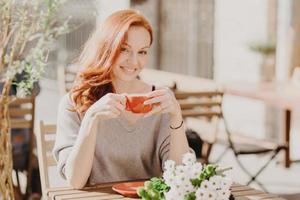 photo horizontale d'une femme heureuse aux cheveux rouges tenant une tasse de boisson, avec une expression heureuse, a des fleurs sur la table, profite d'un rendez-vous avec son petit ami, d'une journée ensoleillée, a une pause-café dans une cafétéria extérieure confortable.