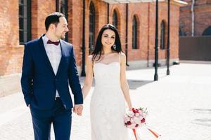 femme brune positive en robe de mariée blanche, tient le bouquet et la main des mariés, célèbre leur mariage ensemble, attend les invités. un couple marié heureux est devenu de jeunes mariés. notion de relation photo