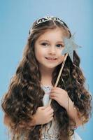 image d'un petit enfant séduisant aux cheveux noirs bouclés, tient une baguette magique, regarde joyeusement au loin, croit au miracle, isolé sur fond bleu. jolie jolie petite fille se tient à l'intérieur photo