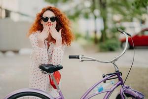 photo en extérieur d'une magnifique femme aux cheveux rouges couvre la bouche avec les deux paumes, a une manucure rouge, vêtue d'une robe à la mode et de lunettes de soleil à la mode, pose près de son vélo, passe du temps libre à faire du vélo