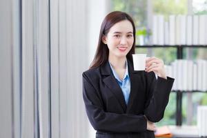 femme d'affaires asiatique qui porte une chemise bleue et un costume noir tient une tasse de café dans sa main et sourit joyeusement au bureau de travail comme arrière-plan. photo