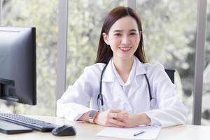 femme médecin professionnelle asiatique assise sourire portant une robe blanche et un stéthoscope en attendant un patient au concept de soins de santé hospitaliers. photo