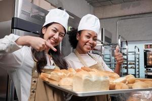 portrait de deux jeunes chefs féminins en uniforme de cuisine regardant la caméra avec un sourire joyeux et fier avec un plateau de pain dans la cuisine. ami et partenaire des aliments de boulangerie, occupation quotidienne de la boulangerie fraîche. photo