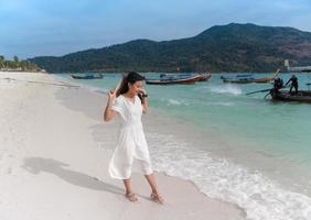 belle femme asiatique en robe blanche tenant un chapeau heureux souriant tout en jouant avec les vagues de la mer sur la plage de sable blanc de la mer bleue photo