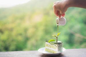 feuille de thé en gros plan et gâteau au thé vert matcha avec poudre de thé vert matcha saupoudré sur le dessus dans une assiette blanche placée sur une table en bois et les mains versent de la sauce au thé vert sur fond vert flou photo