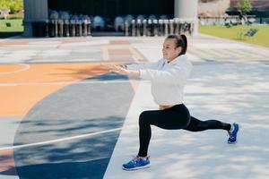 photo horizontale d'une femme sportive motivée qui étire les jambes et les bras fait des exercices d'échauffement avant l'entraînement attend avec impatience vêtue de vêtements de sport pose à l'extérieur sur le grand stade. notion d'exercice