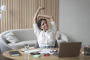 une dame italienne fatiguée employée à distance qui s'étire les bras tout en étant assise au bureau avec un ordinateur portable photo