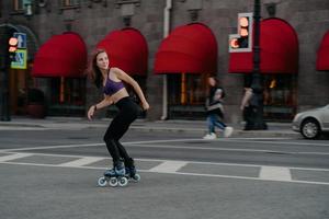 une jeune femme sportive et mince utilise des patins à roulettes comme mode de transport en ville aime son passe-temps préféré et améliore son humeur vêtue de vêtements de sport pose sur la route s'exerce régulièrement garde la forme perd du poids photo