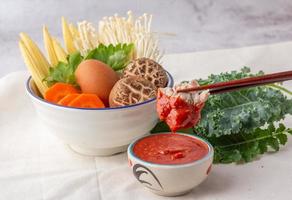 des baguettes de porc cueillies trempées dans un bol de sauce sukiyaki, de nombreux légumes dans un bol blanc comprennent des carottes, du maïs miniature, des champignons shiitake, des aiguilles dorées, du céleri et des œufs de poule. photo