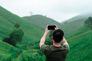 un homme utilisant un téléphone portable prenant une photo d'une montagne verdoyante avec du brouillard le matin.