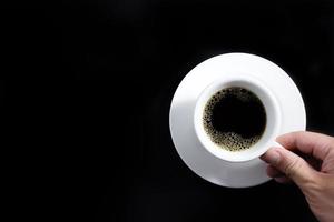 tasse à café noire avec des bulles et un sous-verre avec la main droite tenant la tasse sur un fond noir. photo