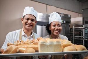 portrait de chefs professionnels en uniforme blanc regardant la caméra avec un sourire joyeux et fier avec un plateau de pain dans la cuisine. un ami et partenaire des aliments de boulangerie et de l'occupation quotidienne de la boulangerie fraîche. photo