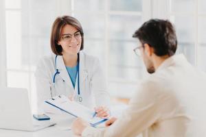 une jeune femme médecin consulte une patiente, affiche des informations et une ordonnance dans le presse-papiers, parle d'un examen médical, pose à la clinique, explique le contrat d'assurance maladie, lit le contrat de service. photo