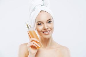 jeune femme au sourire positif, tient une grande bouteille de parfum aromatique à l'odeur de fleur, a un teint soigné, une peau saine, porte une serviette de bain sur la tête, se tient torse nu isolé sur fond blanc photo