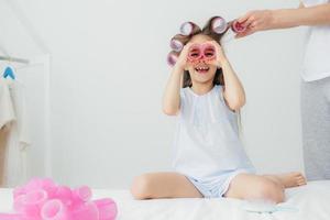 une enfant de sexe féminin mignonne et heureuse s'amuse, garde deux bigoudis près des yeux, porte des vêtements de nuit, sa mère enroule des bigoudis sur ses longs cheveux, pose dans une salle blanche. concept d'enfants, de beauté, de bonheur et de joie photo