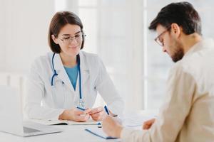 le patient masculin écrit le formulaire de demande, obtient des conseils et des conseils sur le traitement à domicile de la part du médecin, utilise un ordinateur portable et un document médical au bureau, pose ensemble au bureau. rendez-vous médecin homme photo