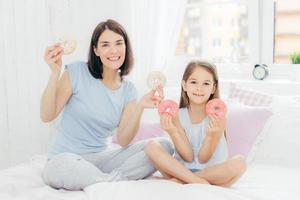 photo horizontale d'une belle femme brune avec une coiffure bobbée, vêtue d'un pyjama, assise près de sa petite fille, tenant ensemble des beignets, allant prendre son petit déjeuner au lit, a d'agréables sourires