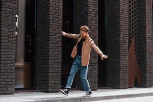 s'amuser en marchant sur le trottoir. homme élégant avec barbe en veste de couleur kaki et en jeans est à l'extérieur près du bâtiment photo