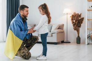 avec le drapeau ukrainien. soldat en uniforme est à la maison avec sa petite fille photo