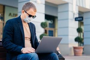 un homme d'affaires concentré pose à l'extérieur près de la gare routière, utilise un ordinateur portable pour le travail à distance, porte un masque de protection pour éviter le coronavirus. concept de personnes, de technologie, de voyage et d'épidémie de virus photo