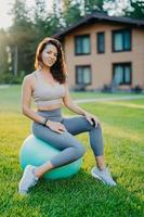 photo verticale d'une belle femme brune assise sur un ballon de fitness sur l'herbe verte et la maison en arrière-plan, vêtue de vêtements de sport, a un entraînement de yoga ou de gymnastique, étant en bonne forme. mode de vie actif