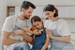 une petite fille affectueuse embrasse un chien de race avec amour, mère et père posent près, passent du temps libre à la cuisine, préparent un délicieux petit déjeuner. famille heureuse et leur animal de compagnie s'aiment beaucoup photo