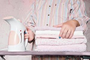 vue en coupe d'une femme tenant un cuiseur à vapeur électrique en tissu et une pile de serviettes prêtes à être repassées sur le bureau de repassage. Gadgets de routine pour le ménage photo