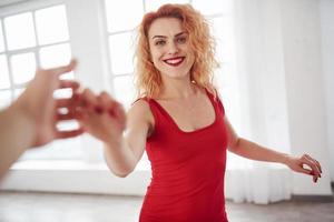 dansons. jolie femme en robe rouge et cheveux bouclés dans la chambre spacieuse près de la fenêtre photo