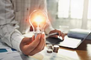 main de femme d'affaires tenant une ampoule avec pile de pièces sur le bureau. concept d'économie d'énergie photo