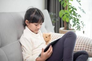 petite fille utilisant une tablette jouant à un jeu sur Internet, enfant assis sur un canapé regardant ou parlant avec un ami en ligne, enfant se relaxant dans le salon le matin, enfants avec un nouveau concept technologique photo