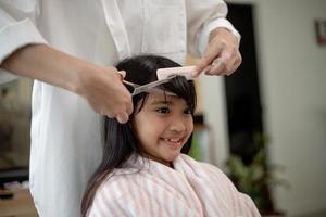 mère asiatique coupant les cheveux à sa fille dans le salon à la maison tout en restant à la maison à l'abri du coronavirus covid-19 pendant le verrouillage. concept d'auto-quarantaine et de distanciation sociale. photo