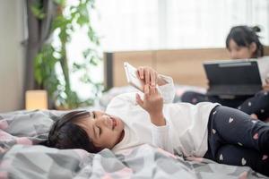 jolie petite fille regardant une vidéo sur un smartphone avec un visage souriant seul sur le lit, enfant utilisant un téléphone portable avec un visage heureux à la maison. rester à la maison quarantaine coronavirus covid-19 prévention de la pandémie. photo