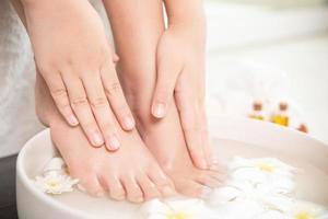 vue rapprochée d'une femme trempant sa main et ses pieds dans un plat avec de l'eau et des fleurs sur un plancher en bois. traitement de spa et produit pour les pieds féminins et le spa des mains. fleurs blanches dans un bol en céramique. photo