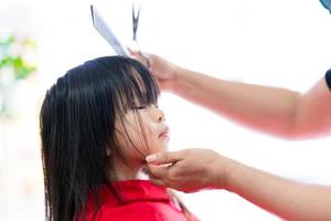portrait d'enfant de 3-4 ans. jolie fille asiatique reste immobile pour que le coiffeur coiffe magnifiquement les cheveux. les mains des esthéticiennes tiennent un peigne et des ciseaux et soutiennent doucement son petit visage. photo