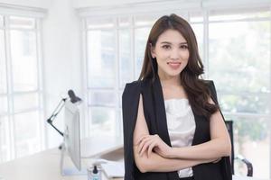 une travailleuse asiatique qui porte un costume noir et des bras croisés se tient dans une salle de bureau blanche, a l'air intelligente et a un ordinateur et un bureau en arrière-plan. photo