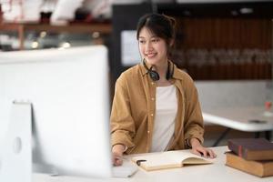 hipster fille asiatique écolière avec porter casque écrire des notes regarder la vidéo en ligne webinaire apprendre sur ordinateur portable s'asseoir dans son lit cours d'apprentissage en ligne à distance vidéoconférence pc appeler dans la chambre à la maison photo