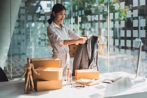 jeune créatrice de mode asiatique utilisant un ordinateur recevant un bon de commande et montrant des vêtements enregistrant une vidéo en direct en ligne à la maison. propriétaire de petite entreprise, concept de livraison de marché en ligne. photo