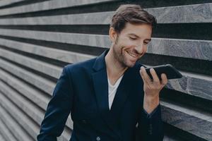 jeune homme séduisant se tient à l'extérieur et utilise un assistant de reconnaissance vocale sur smartphone photo