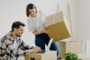 un couple familial fraîchement marié pose dans leur nouvel appartement, déballe des cartons avec des effets personnels, un homme heureux est assis devant un ordinateur portable, recherche des idées de conception, a des problèmes de routine domestique photo