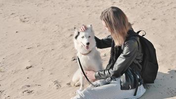 jeune femme portant une veste et une robe assise sur un sable à côté de son chien. laika yakoutienne blanche et son propriétaire passent du temps et se détendent ensemble sur une plage photo