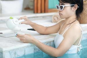 femme heureuse en maillot de bain blanc buvant du thé chaud dans un hôtel de luxe avec piscine, jeune femme avec chapeau profiter d'un complexe tropical. concept de détente, de voyage d'été, de vacances, de vacances et de week-end photo