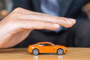 couverture de main d'homme d'affaires ou jouet de voiture orange de protection sur la table. concept financier, monétaire, de refinancement et d'assurance automobile photo