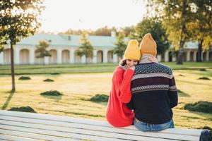 belle femme en pull rouge lâche embrasse son petit ami, lui a trop manqué comme je ne l'ai pas vu il y a longtemps, s'asseoir sur un banc dans le parc, avoir une conversation agréable, admirer le magnifique paysage ou la nature photo